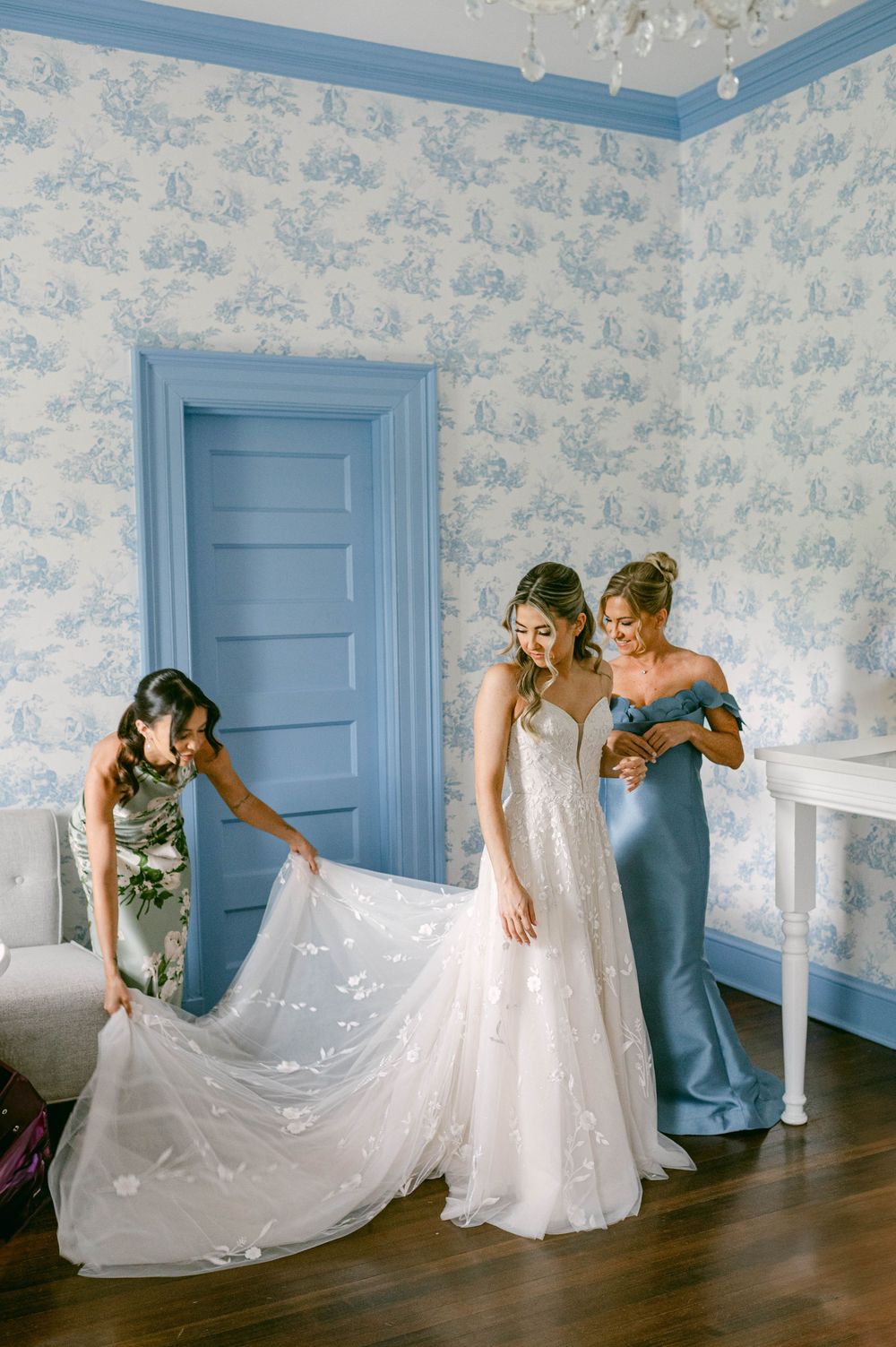 Wedding party helps adjust a flowing white wedding gown in a room with blue and white floral wallpaper.