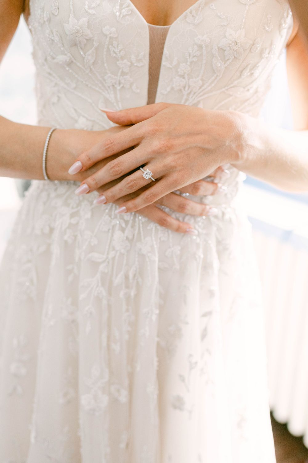 Close-up of hands wearing delicate bridal jewelry against a white lace wedding dress with intricate floral details.