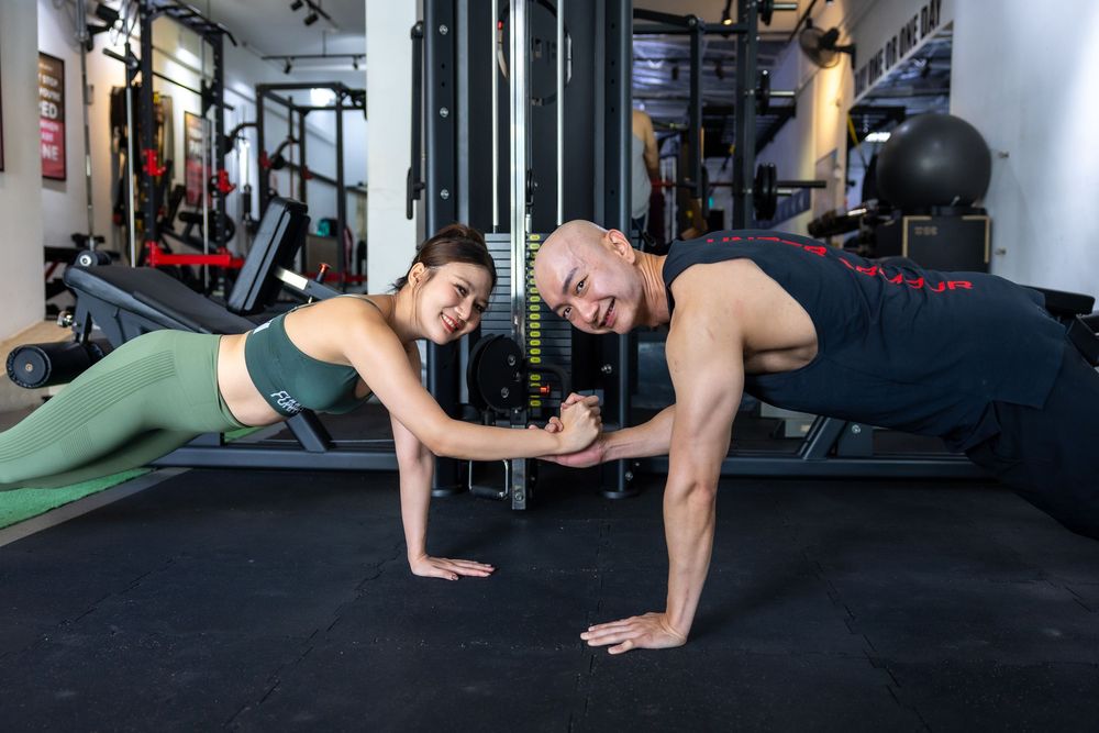 Two people performing plank exercises together in a well-equipped fitness gym with workout equipment in background.