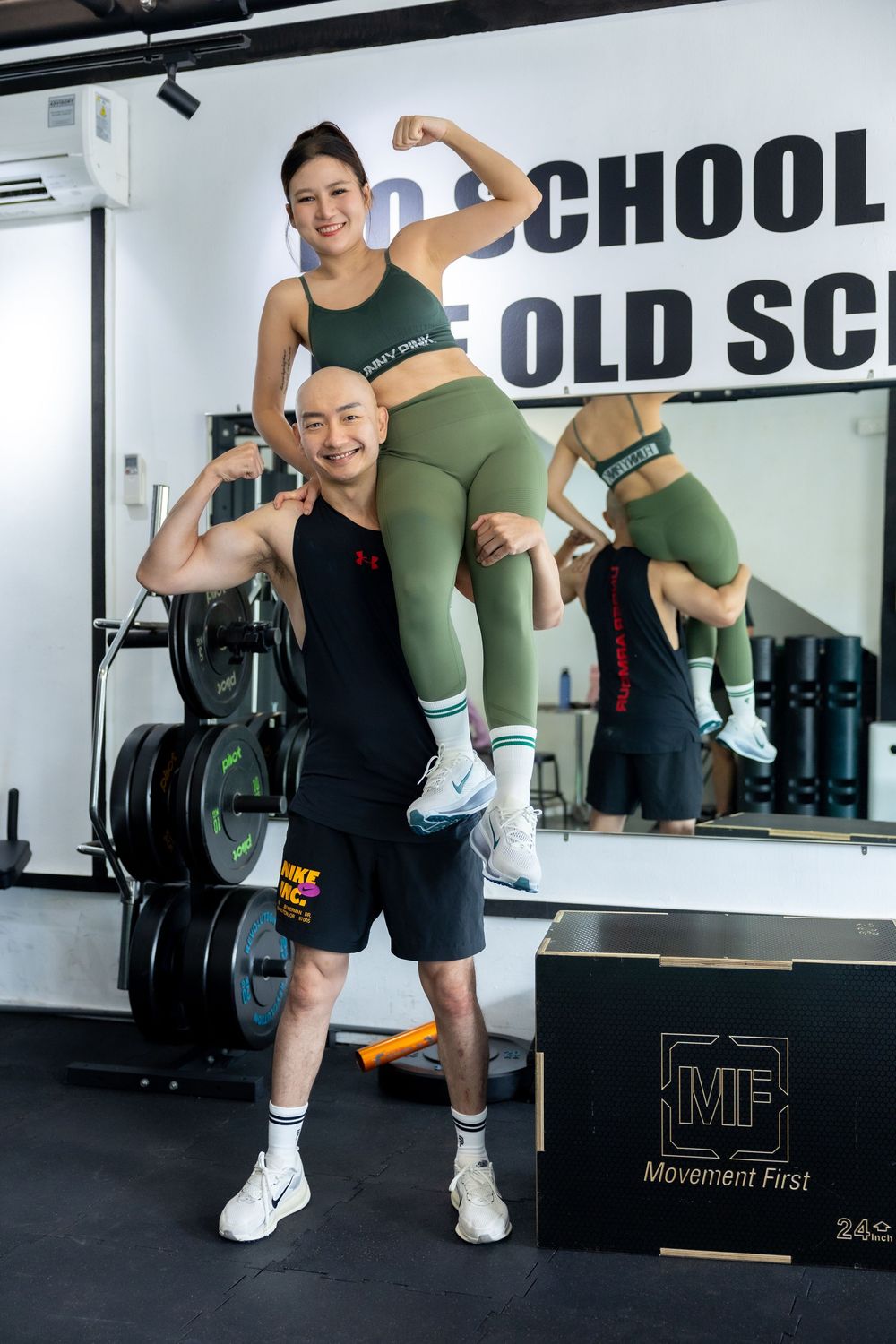 Two people pose playfully at a fitness gym with weight equipment and motivational wall text visible in the background.