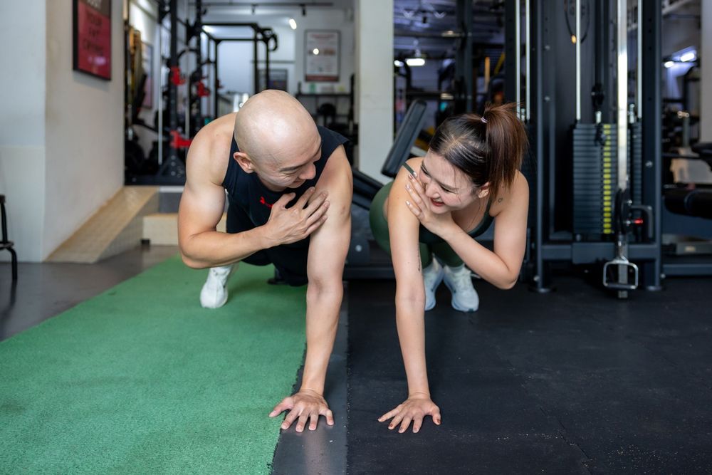 Two people doing plank exercise together on a green mat in a gym setting while smiling at each other.