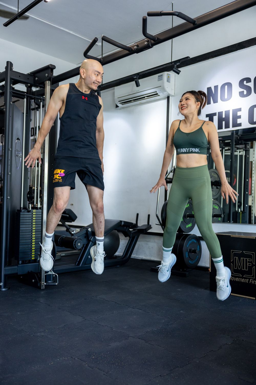 Two people exercising together in a modern gym, doing synchronized jumping movements on a black rubber floor.