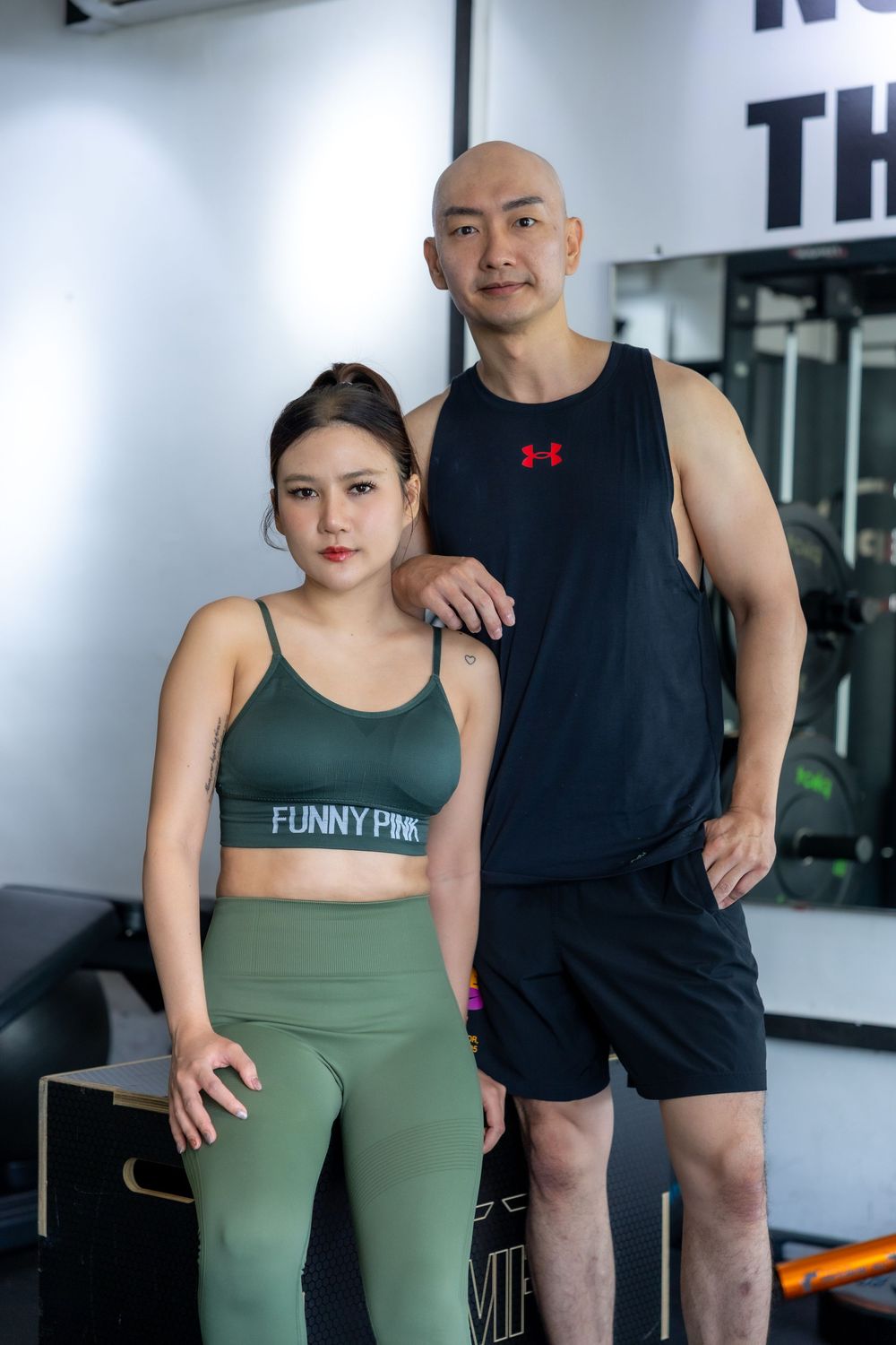 Two fitness enthusiasts pose together at a gym, wearing athletic wear including a green sports bra and black tank top.