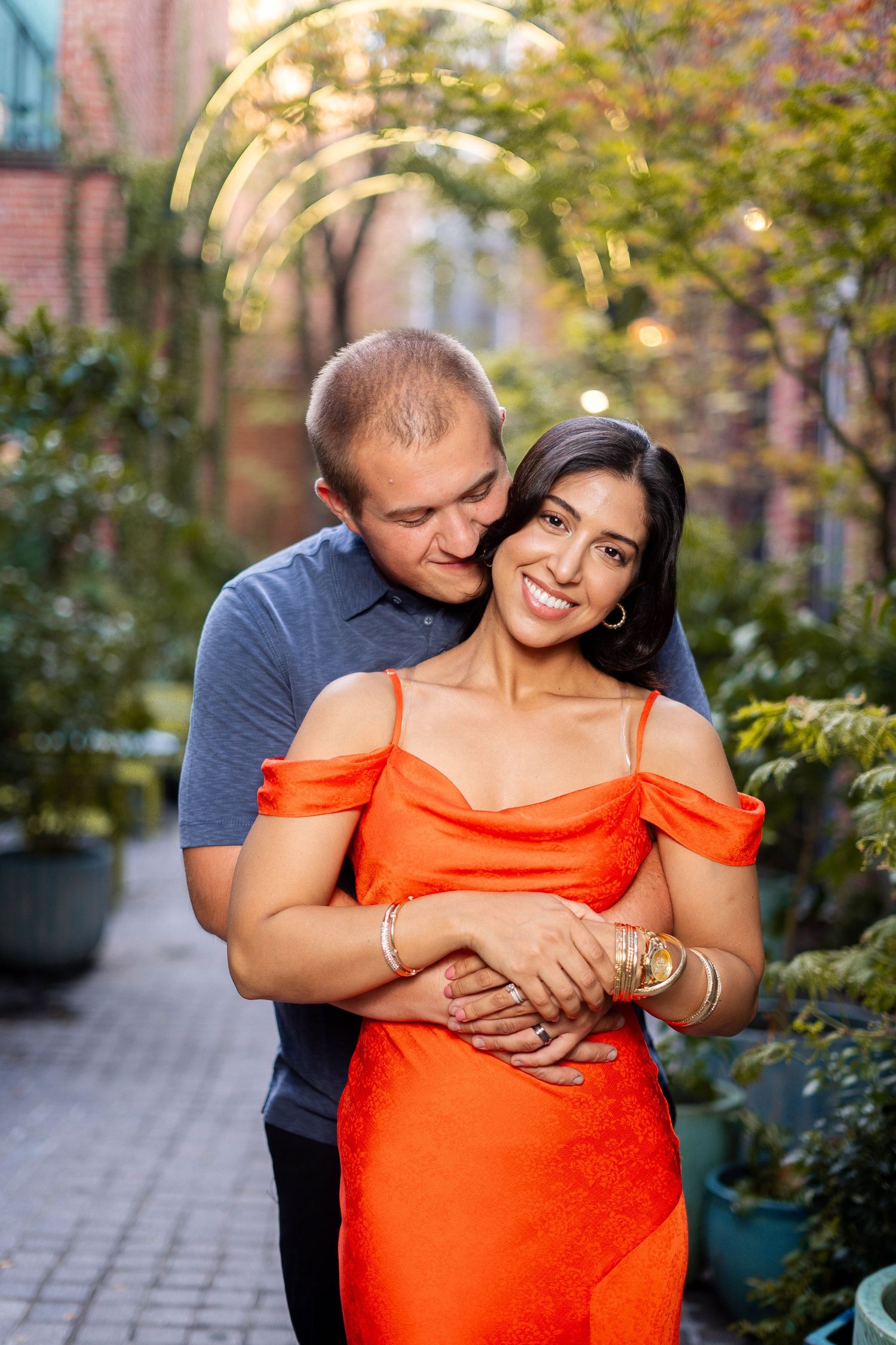 Smiling couple embracing in a garden, woman wearing an orange off-shoulder dress.