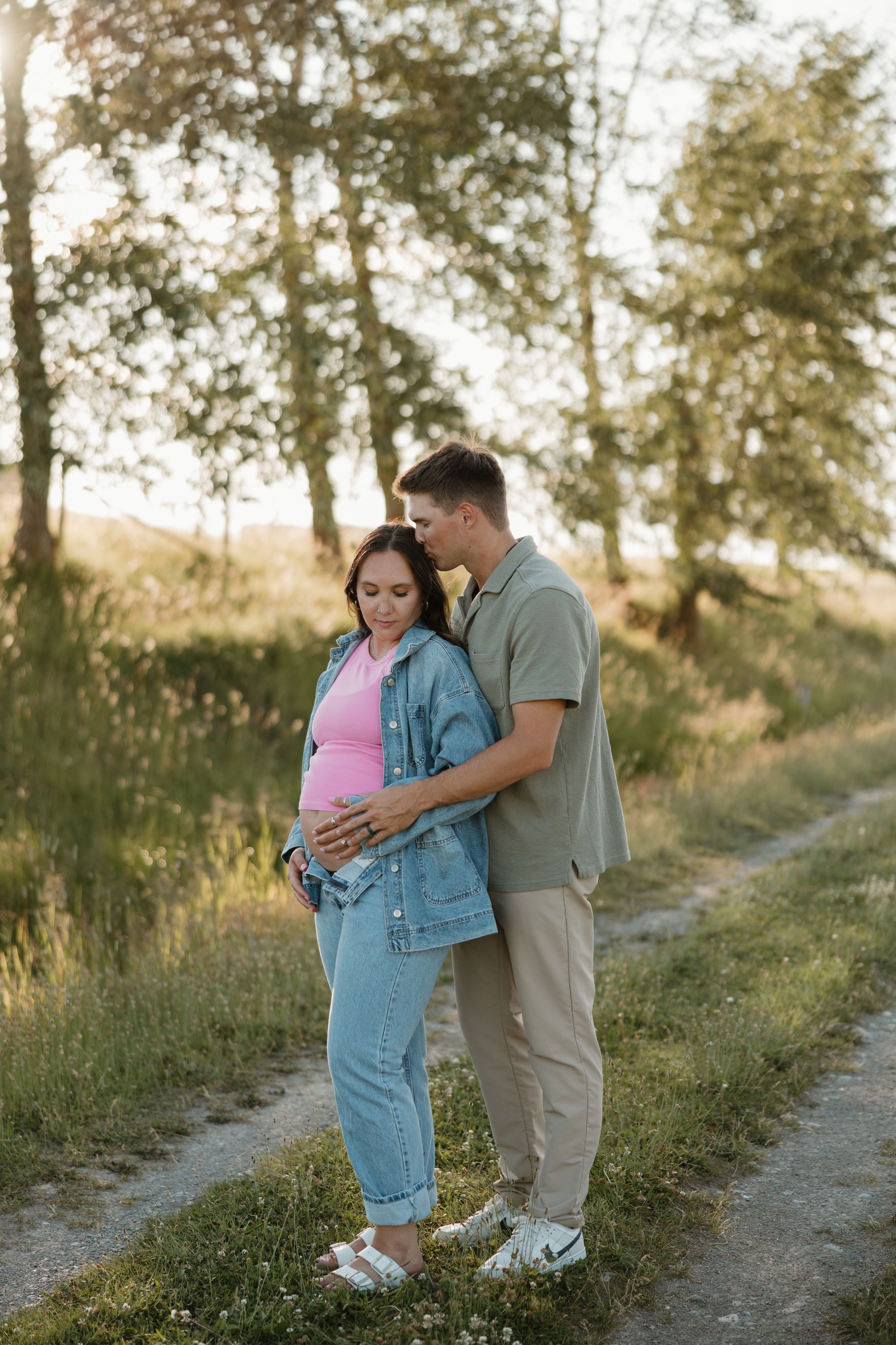 A couple embraces on a sunlit dirt path among trees and greenery during a romantic maternity photo session.