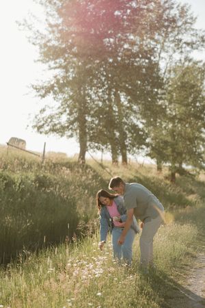 Two people bend down to look at wildflowers along a sunlit hillside path during golden hour.