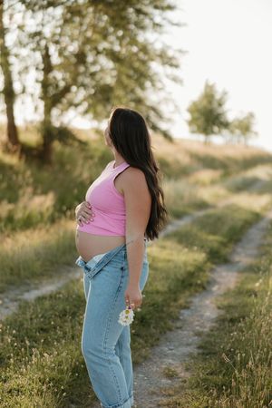 A pregnant woman in a pink tank top and jeans stands on a dirt path during golden hour.