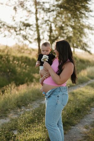 A parent in jeans and pink tank top holds a baby on a dirt path during golden hour in a scenic outdoor setting.