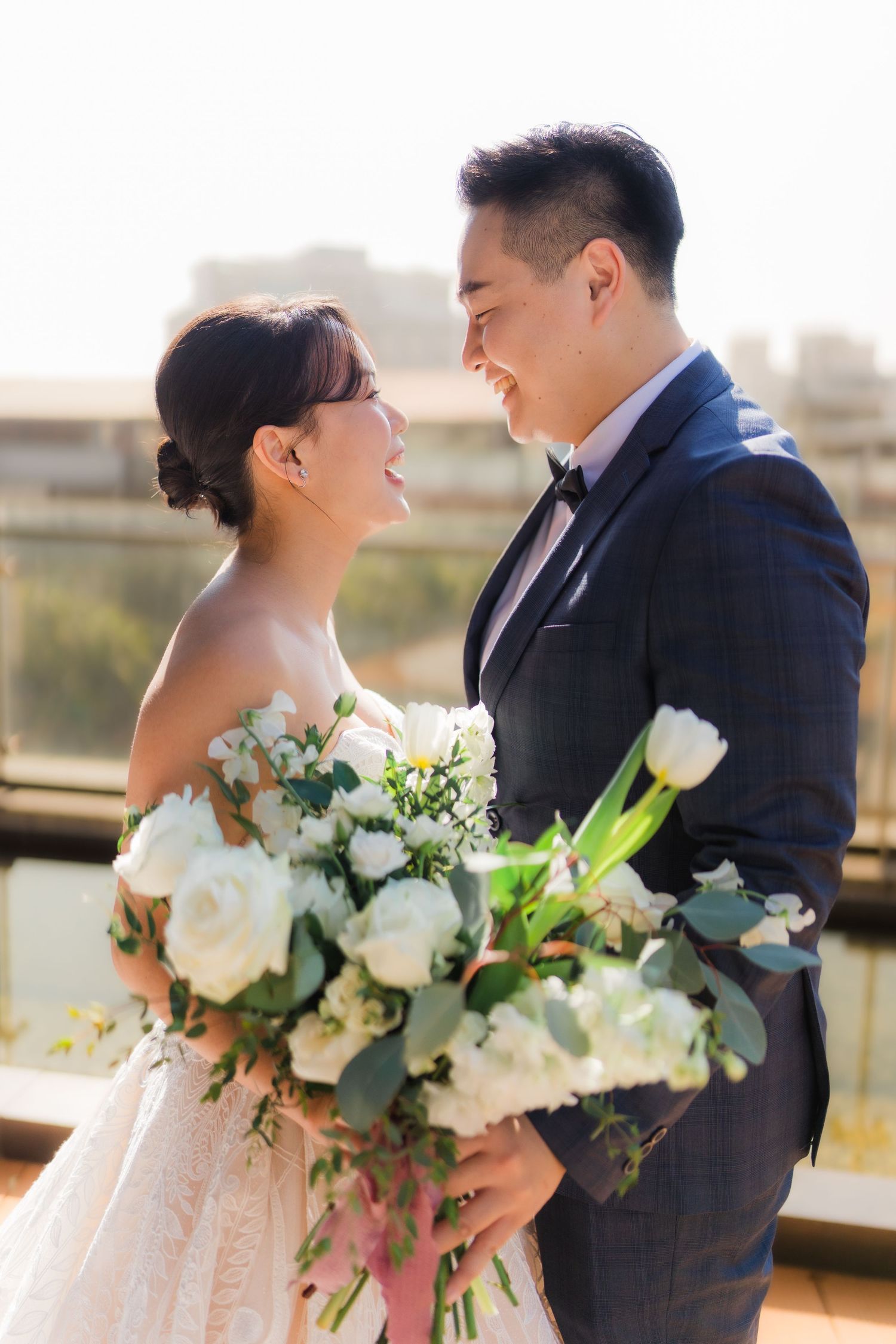 A couple shares an intimate moment on their wedding day with a lush white floral bouquet against a sunlit backdrop.