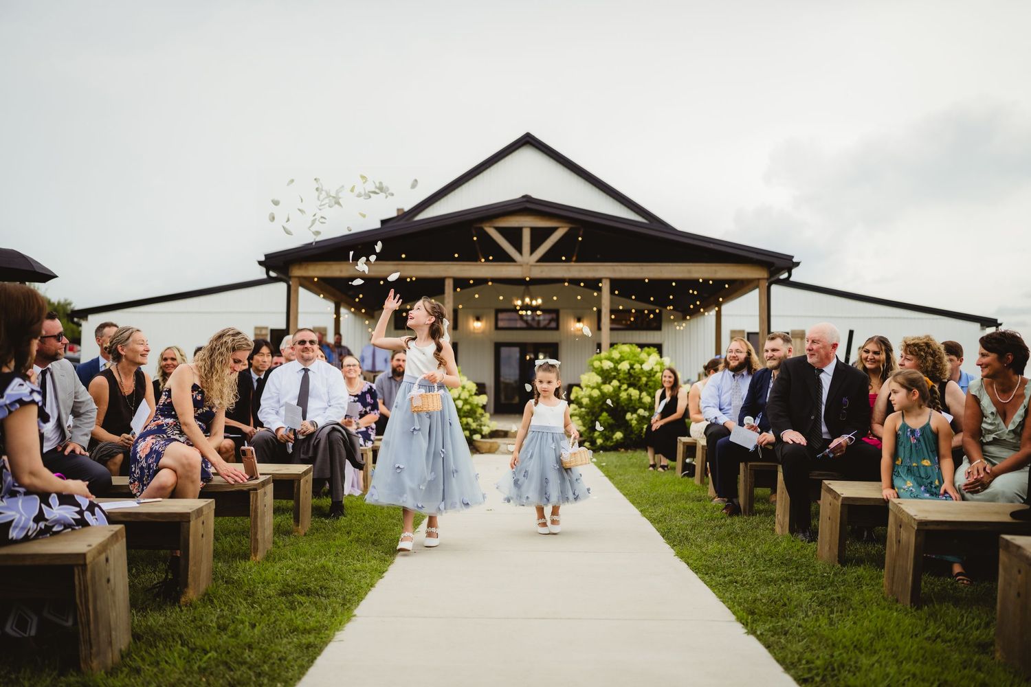 Wedding ceremony takes place on white aisle with rustic wooden benches and crowd of guests at outdoor venue.