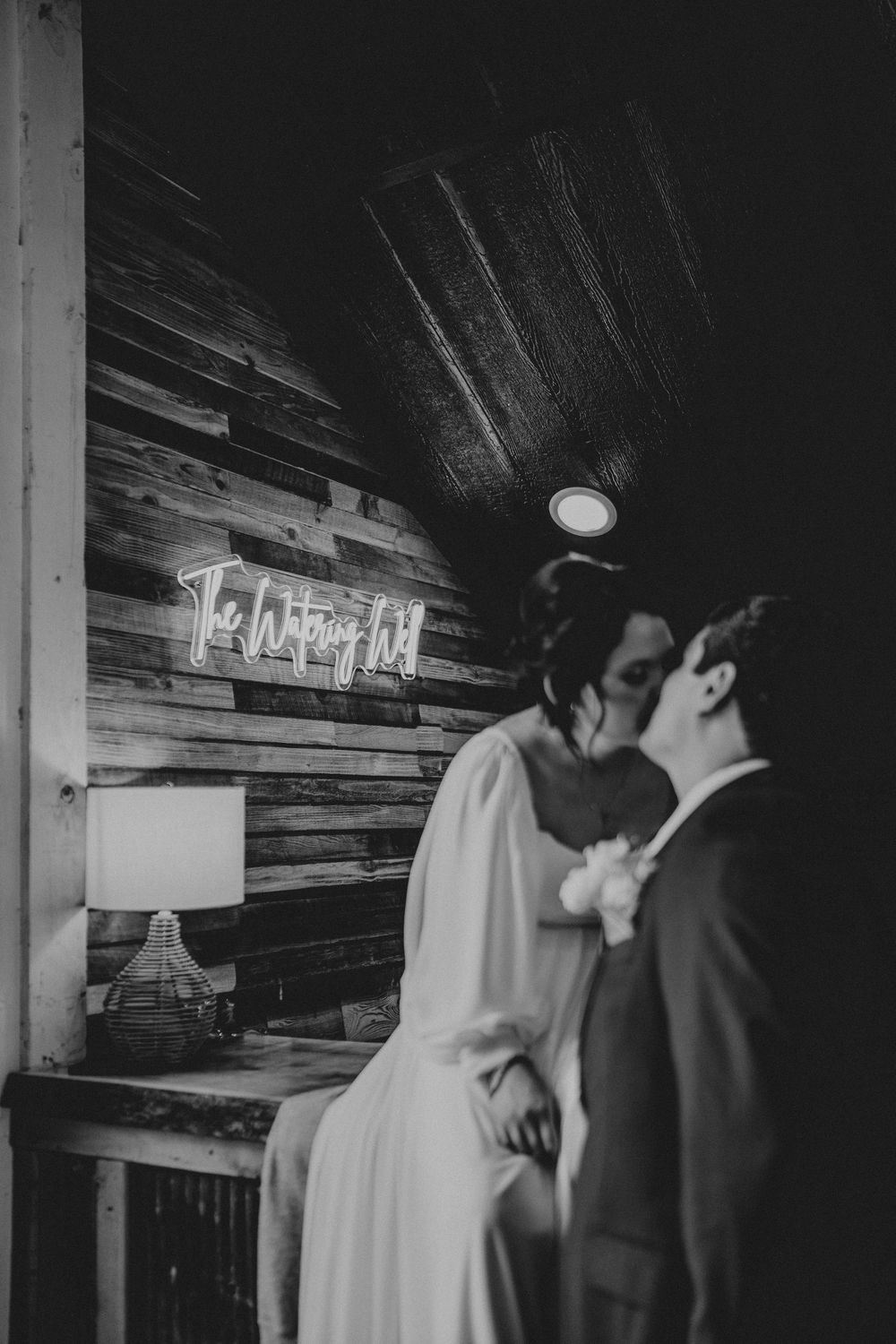 Black and white romantic wedding photo in rustic wood venue with vintage neon sign and table lamp in background.