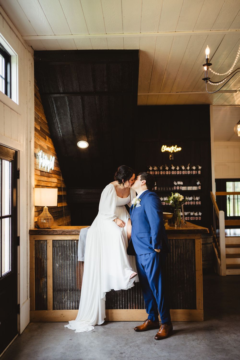 A romantic wedding couple sharing a kiss in a rustic venue with wooden bar and warm lighting in the background.