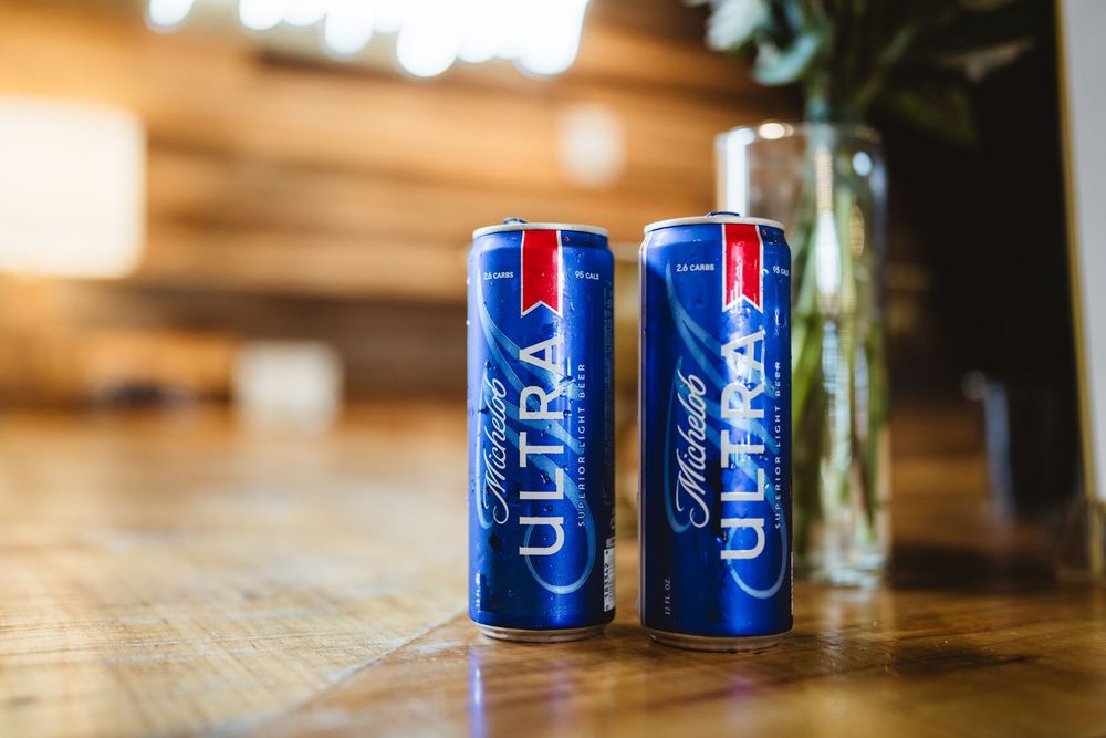 Two blue Michelob Ultra beer cans sit on a wooden surface with soft lighting in the background.