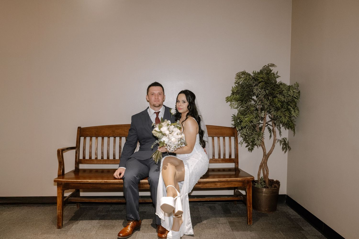 A wedding couple sits together on wooden benches in a minimalist hallway with indoor plants.
