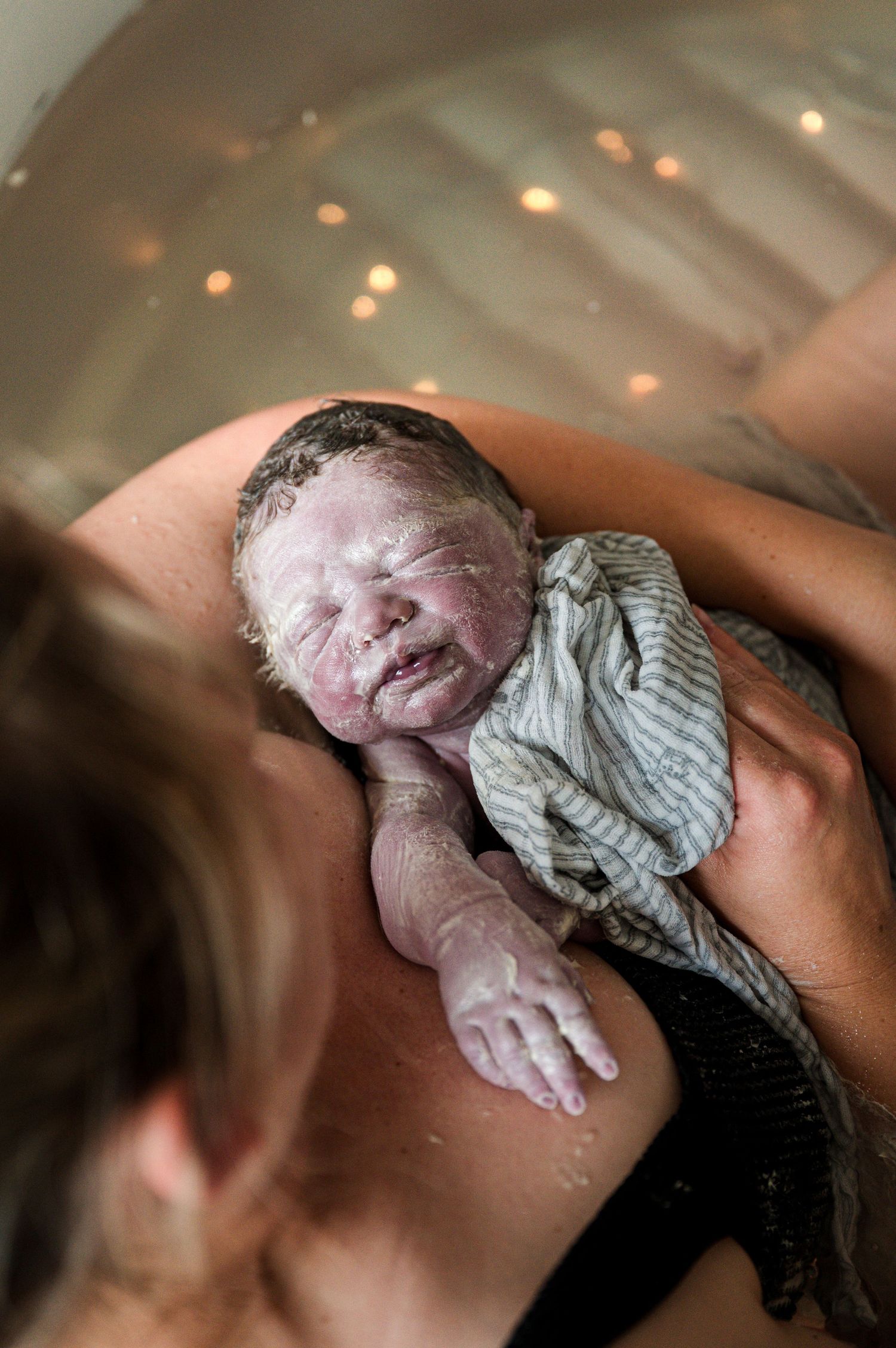 Newborn baby wrapped in striped cloth cries during first moments after birth with bokeh lights in background.