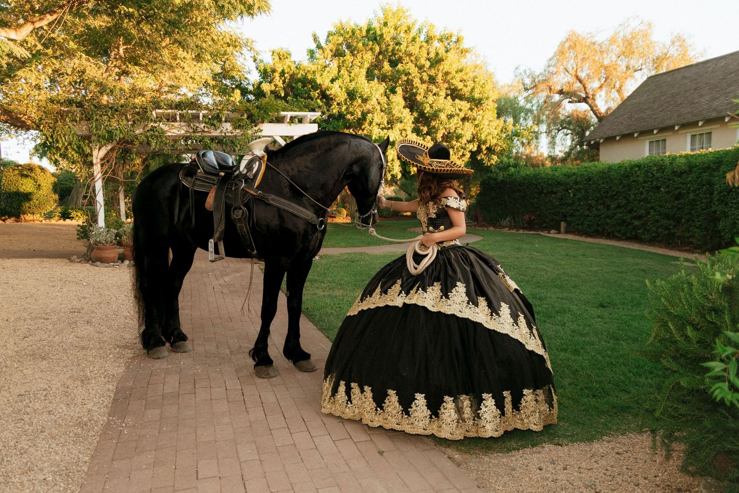 Black horse and figure in an elegant black & gold Quinceañera gown pose on a garden path during golden hour sunset.