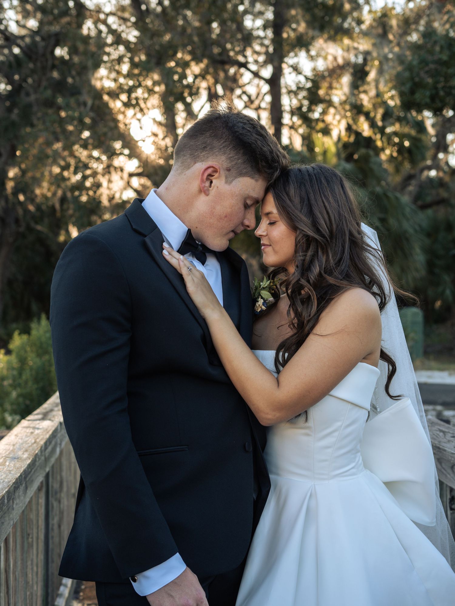 A romantic wedding moment captured at sunset on a wooden bridge as a couple in formal attire embrace tenderly.