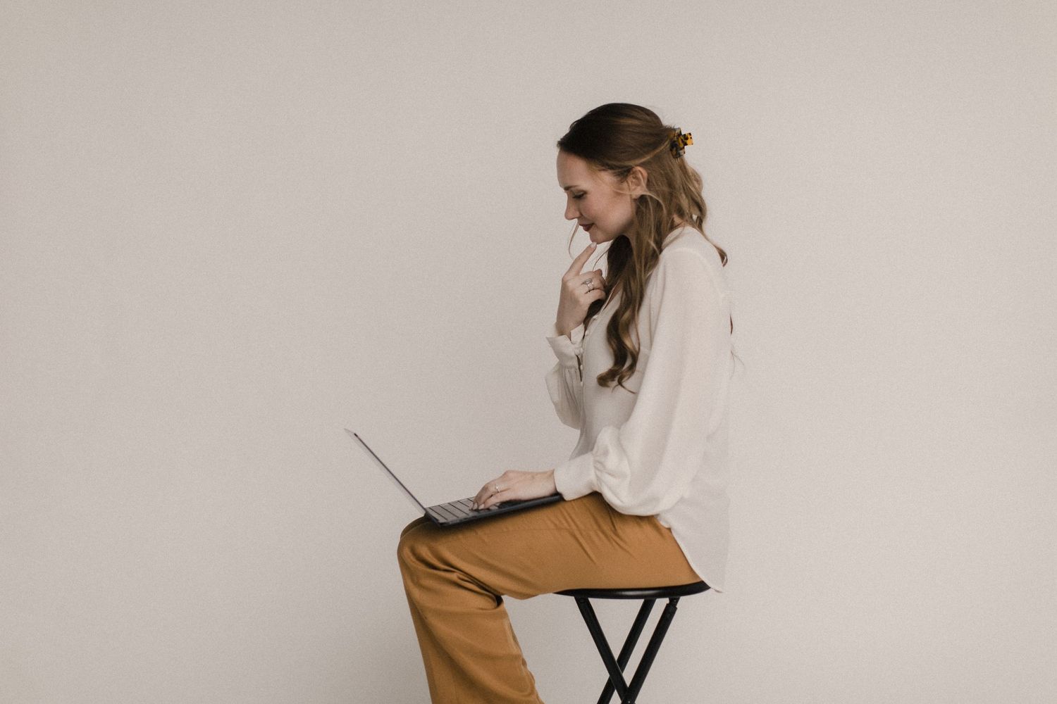 Minimalist scene of person in tan pants and white sweater sitting on a stool while working on laptop.