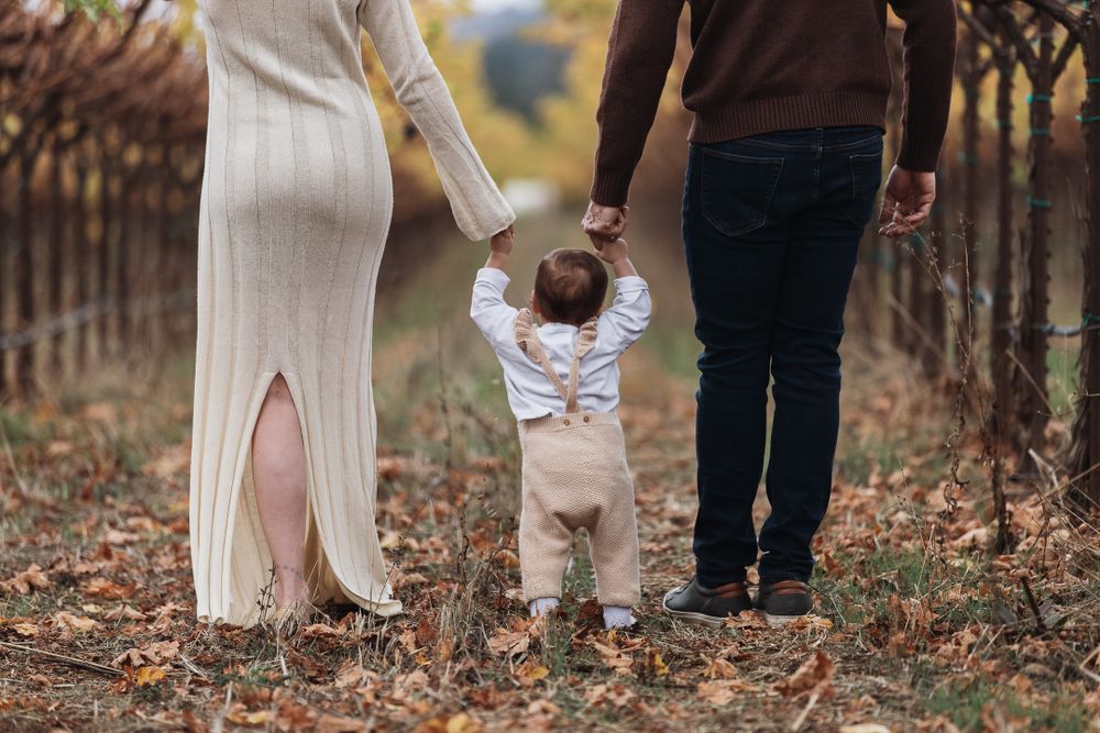 A small child learning to walk holds hands with adults in a fall garden setting among fallen leaves.
