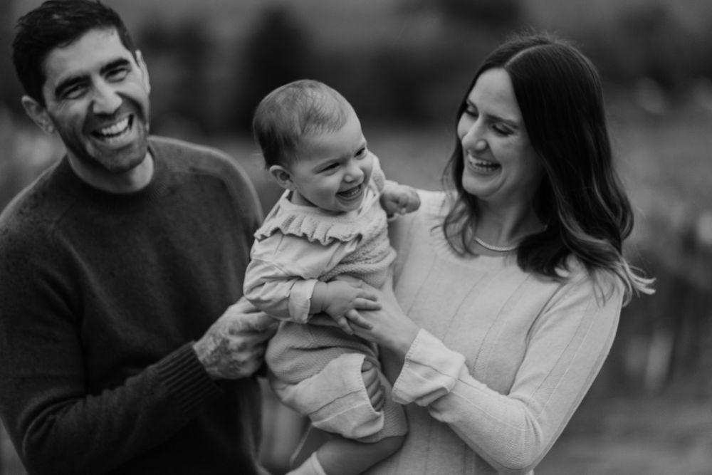 Black and white photo of a happy family laughing together while holding their baby outdoors in casual attire.
