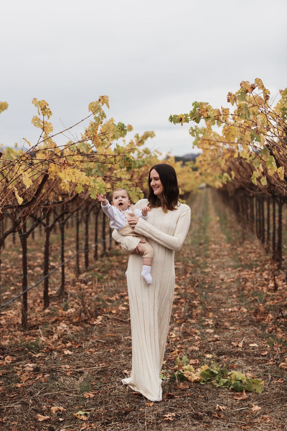 Feminine figure in cream maxi dress stands among autumn vineyard rows holding small baby in serene outdoor setting.