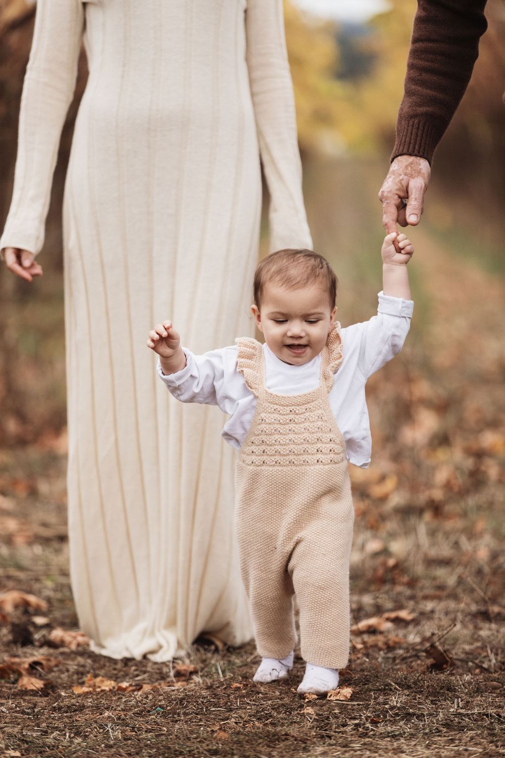 A small child in a beige knit overall takes first steps while holding an adult's hand during an autumn photoshoot outdoors.