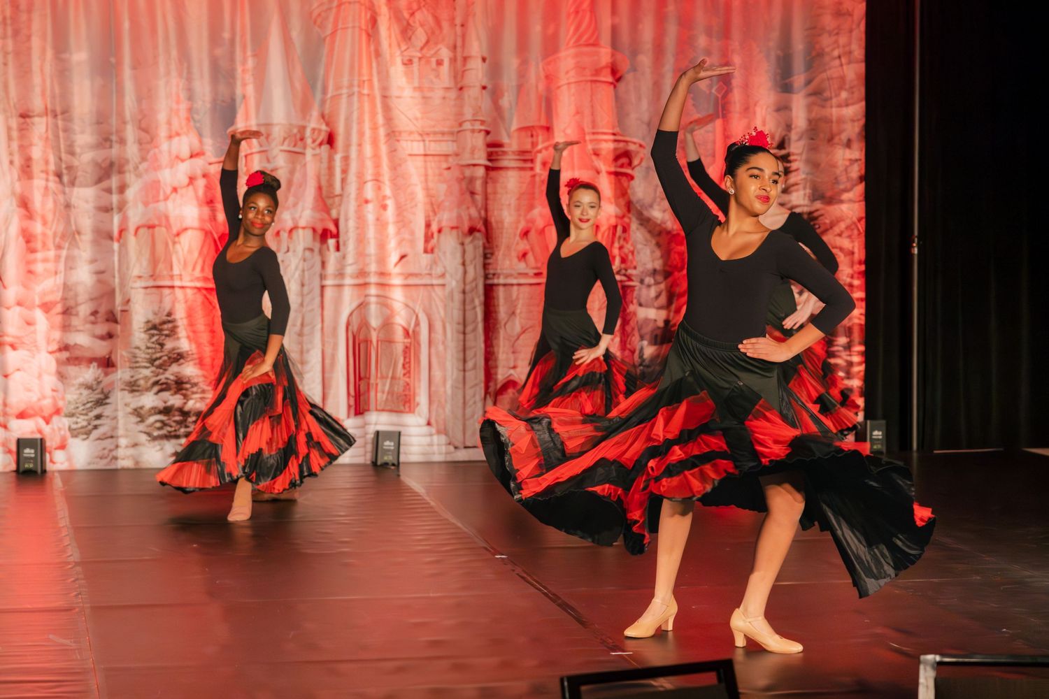 Dancers perform in flowing black and red flamenco dresses under dramatic red stage lighting during a dance performance.