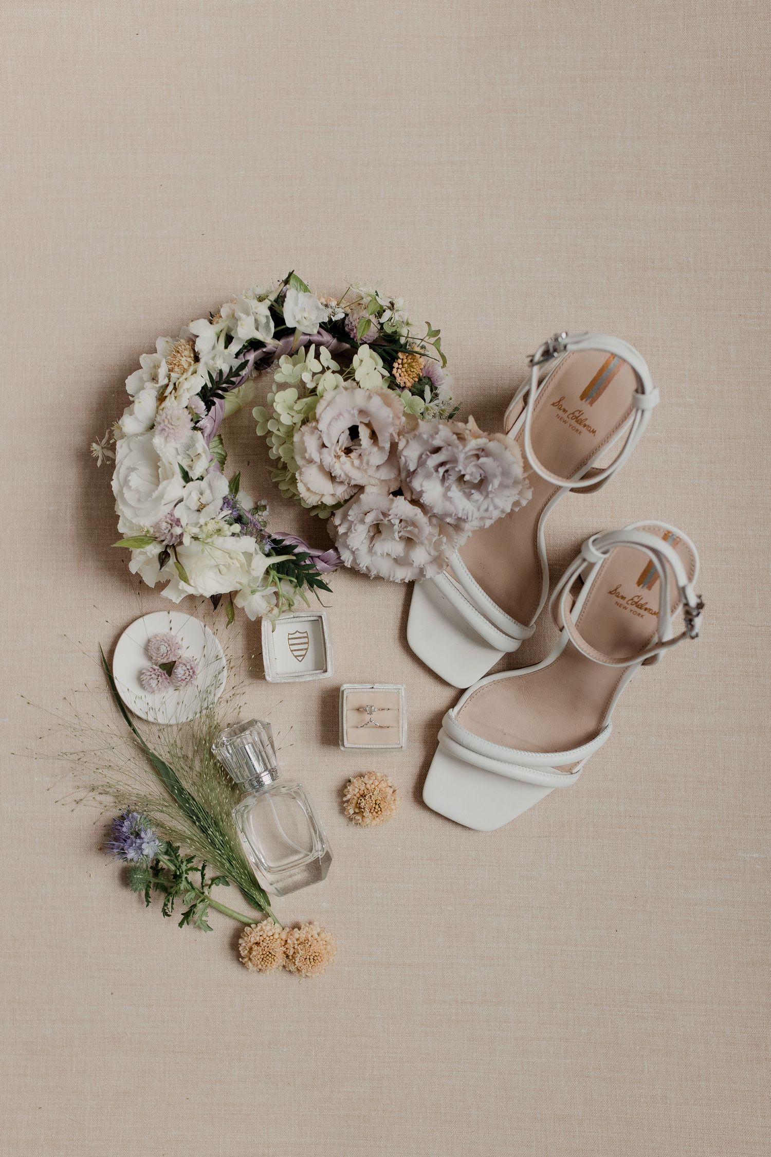 Bridal flat lay with white heeled sandals, floral crown, perfume bottles, and ring box on neutral background.