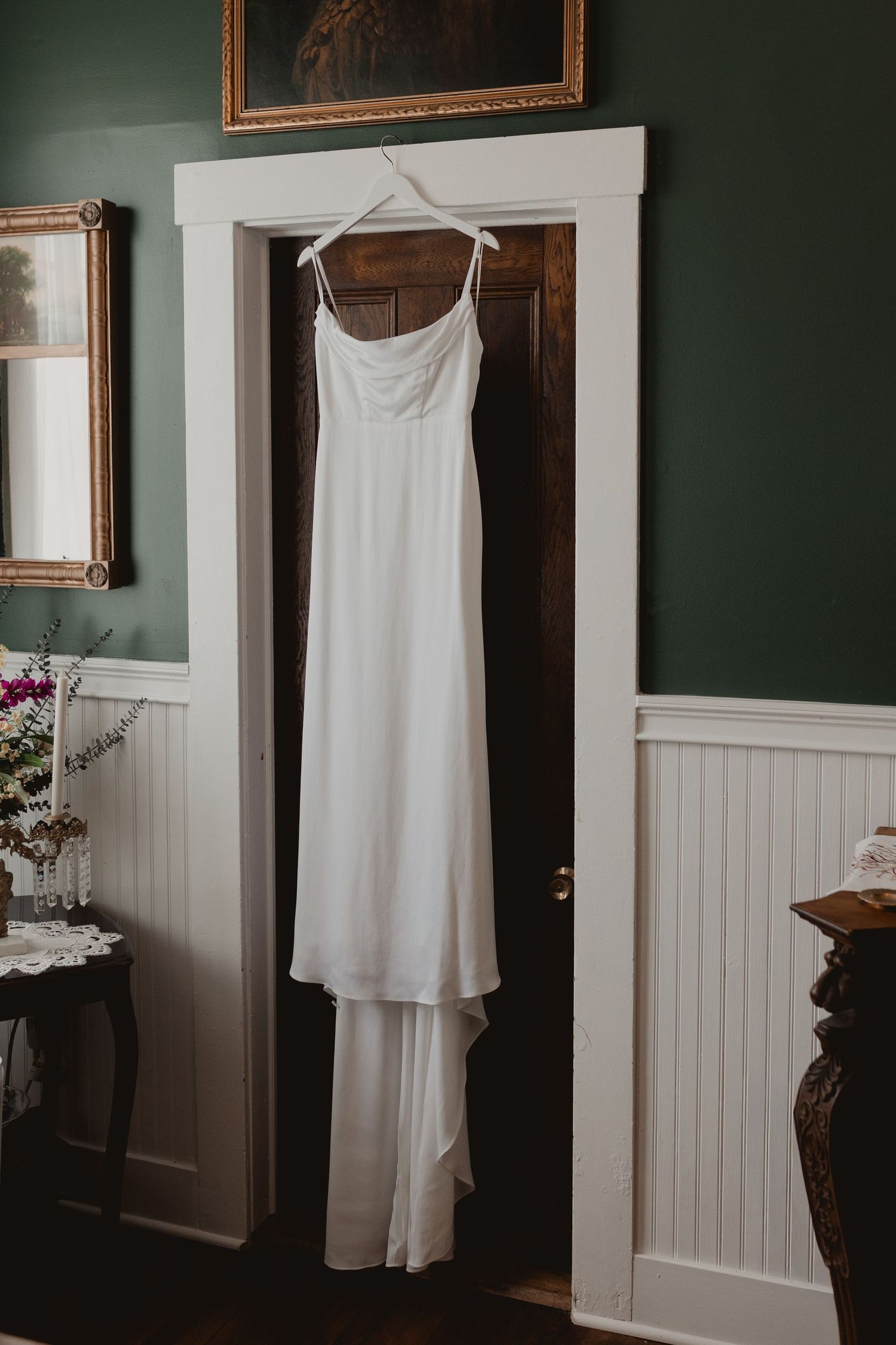 An elegant white wedding dress hanging in a doorway with dark green walls and white wainscoting.