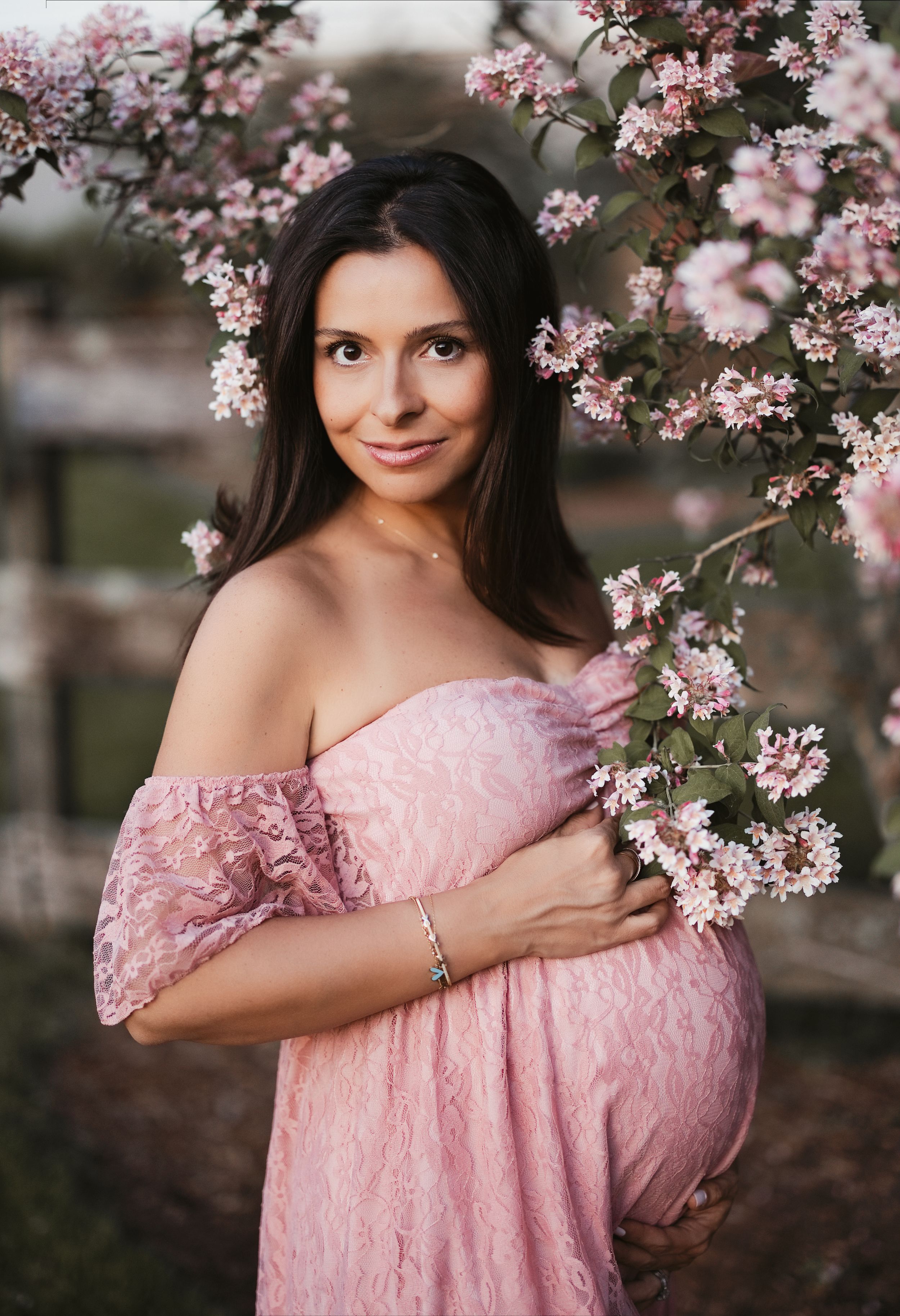 A pregnant woman in an off-shoulder pink lace dress poses gracefully beside blooming pink spring flowers.
