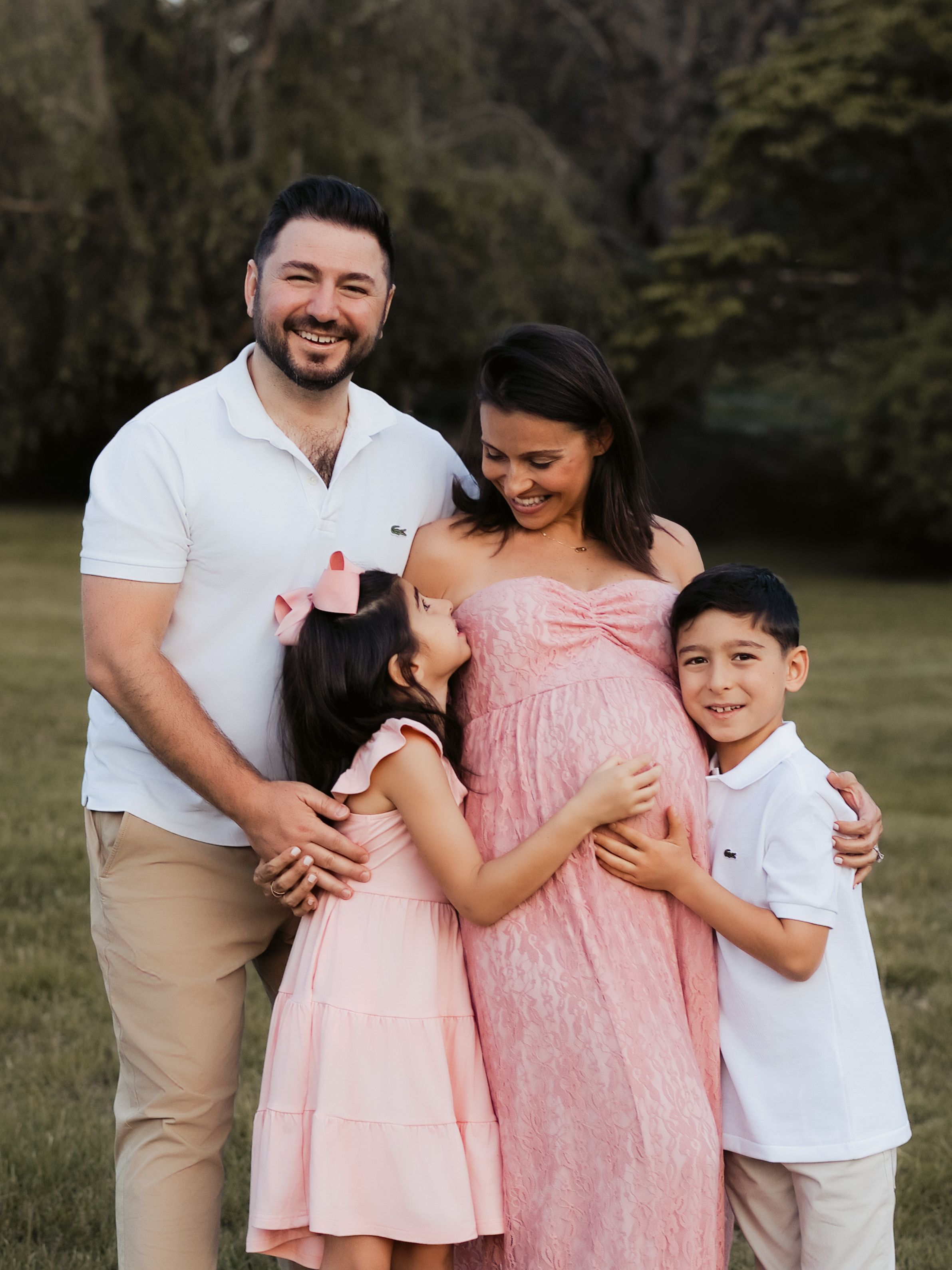 A happy family of four poses outdoors, with two children embracing their smiling pregnant mother in pink.