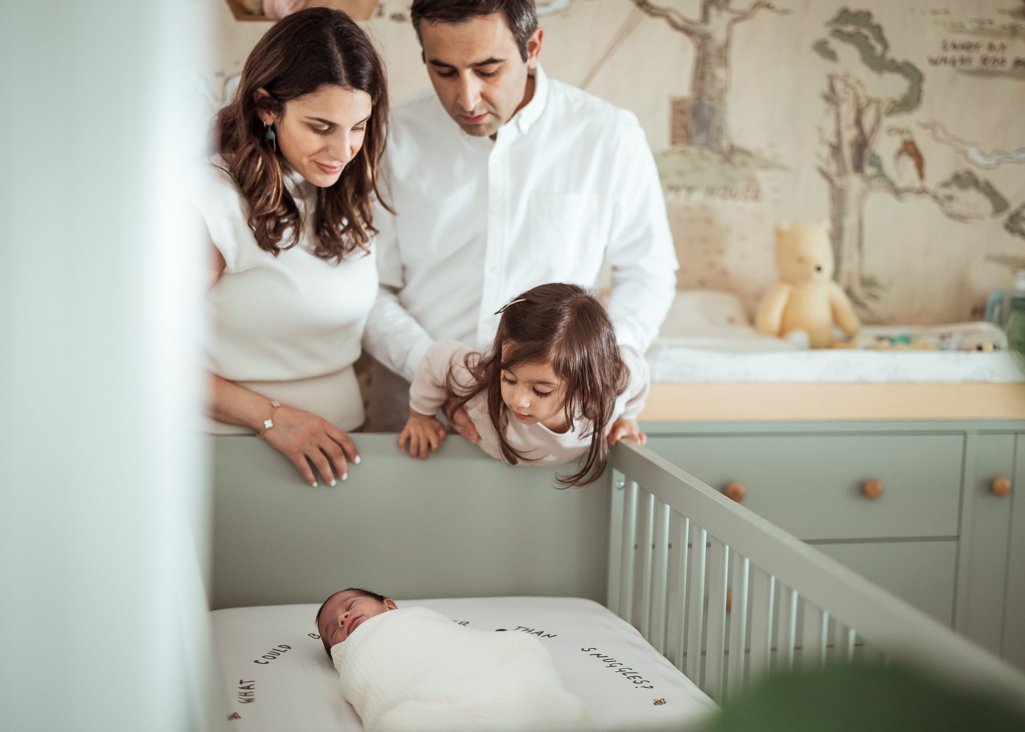 A loving family of four gazes at their swaddled newborn baby resting in a crib in a nursery.