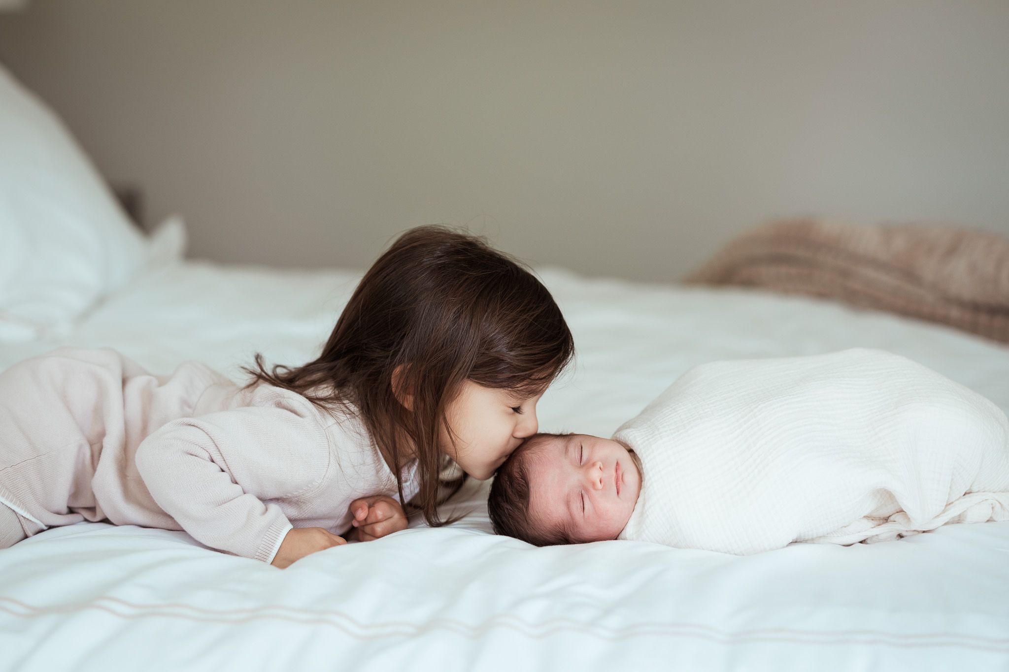 A young toddler girl gently kisses her sleeping swaddled newborn sibling on a white bed.