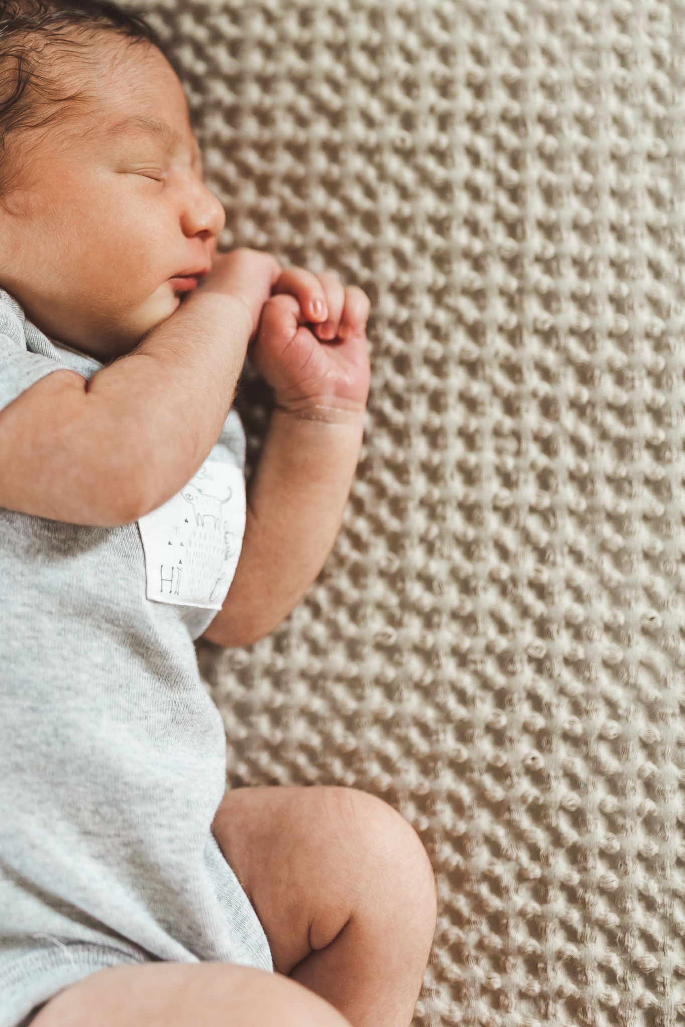 Peaceful newborn baby sleeping on a beige knit blanket in a gray onesie with tiny fists curled near face.