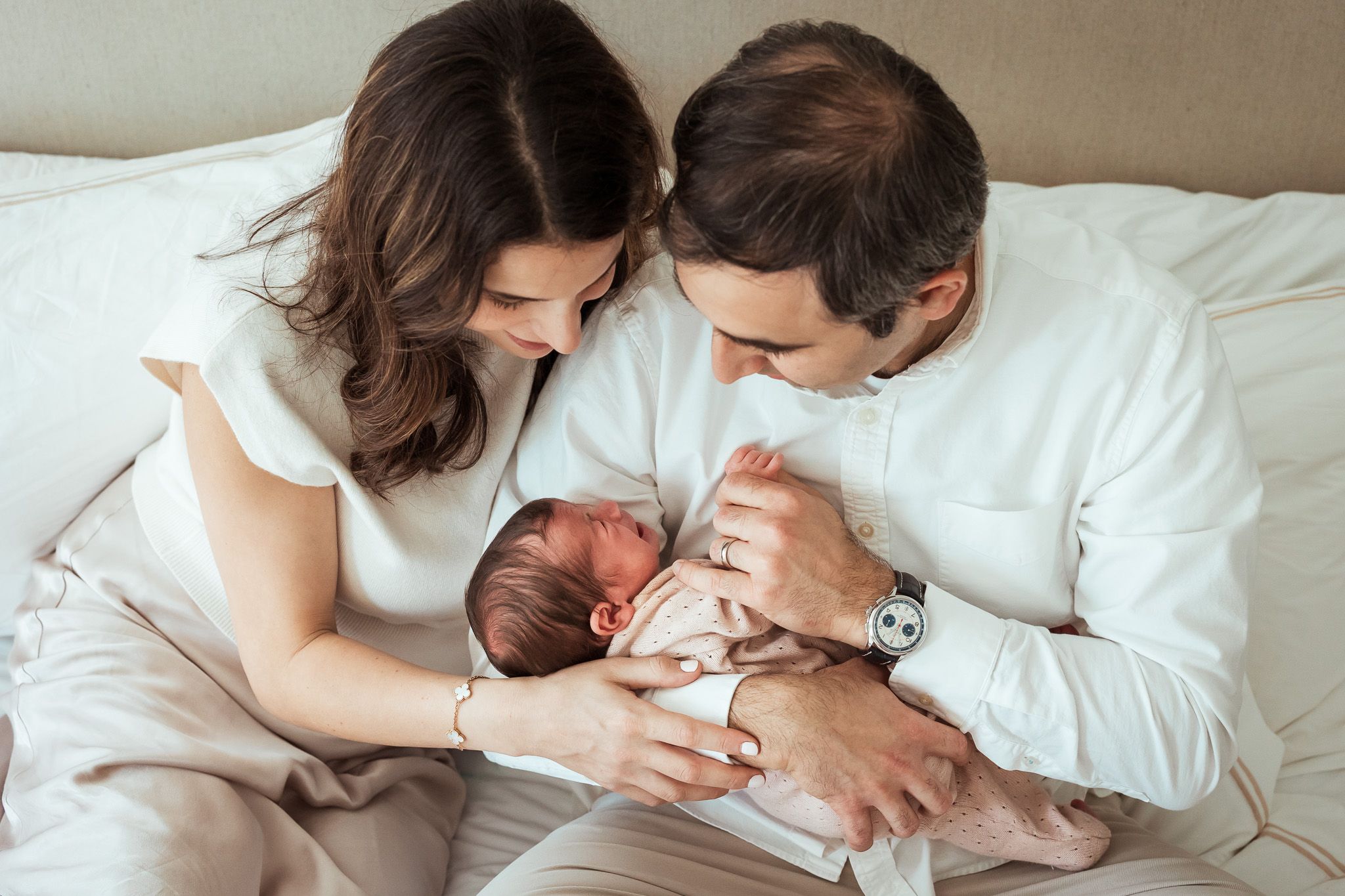 A couple in white clothing lovingly cradles their newborn baby together on a bed during an intimate session.