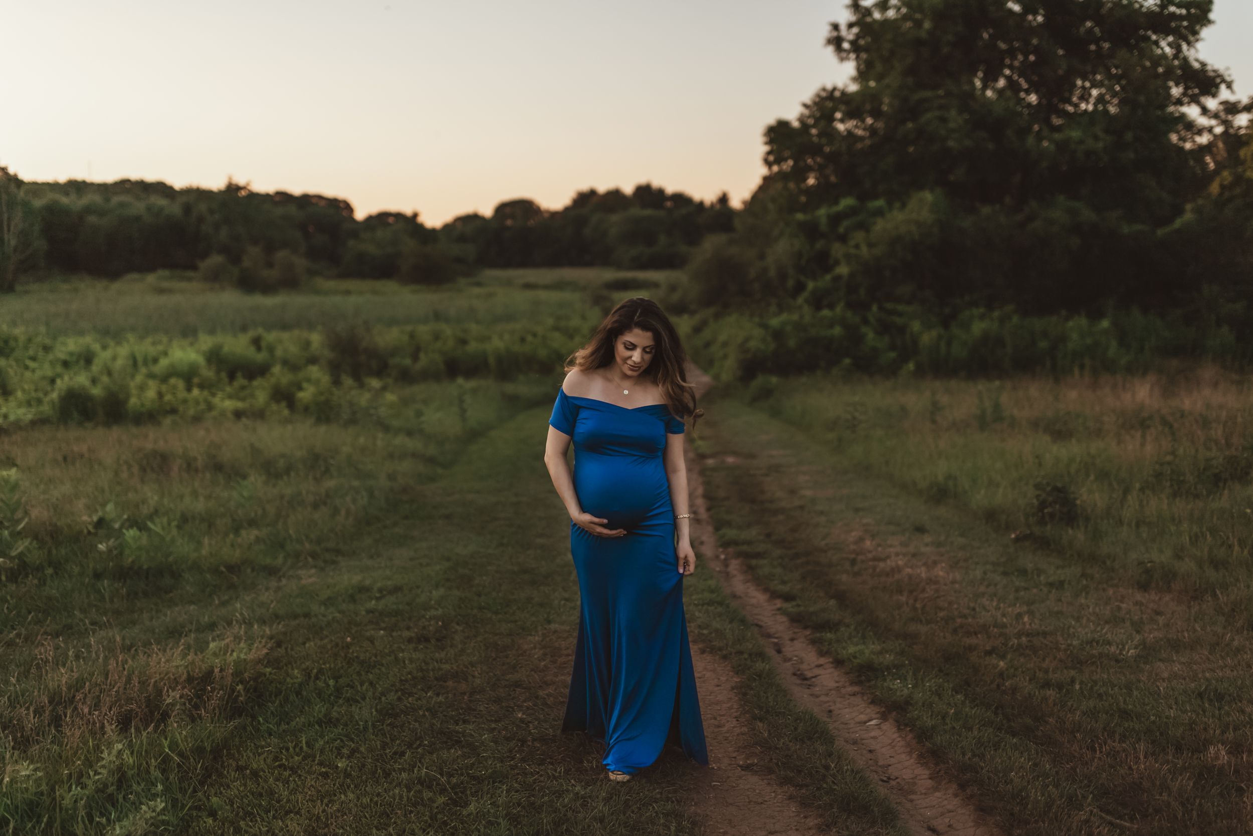 Pregnant woman in blue off-shoulder gown walking on rural dirt path at golden hour sunset.