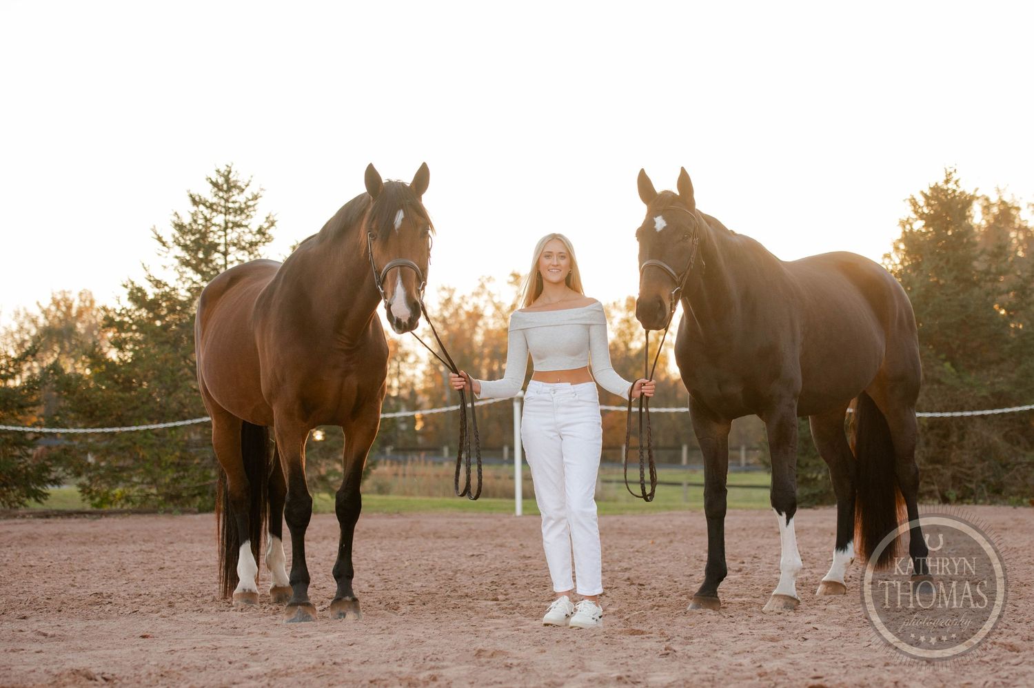 A person stands between two dark horses in a riding arena at sunset.