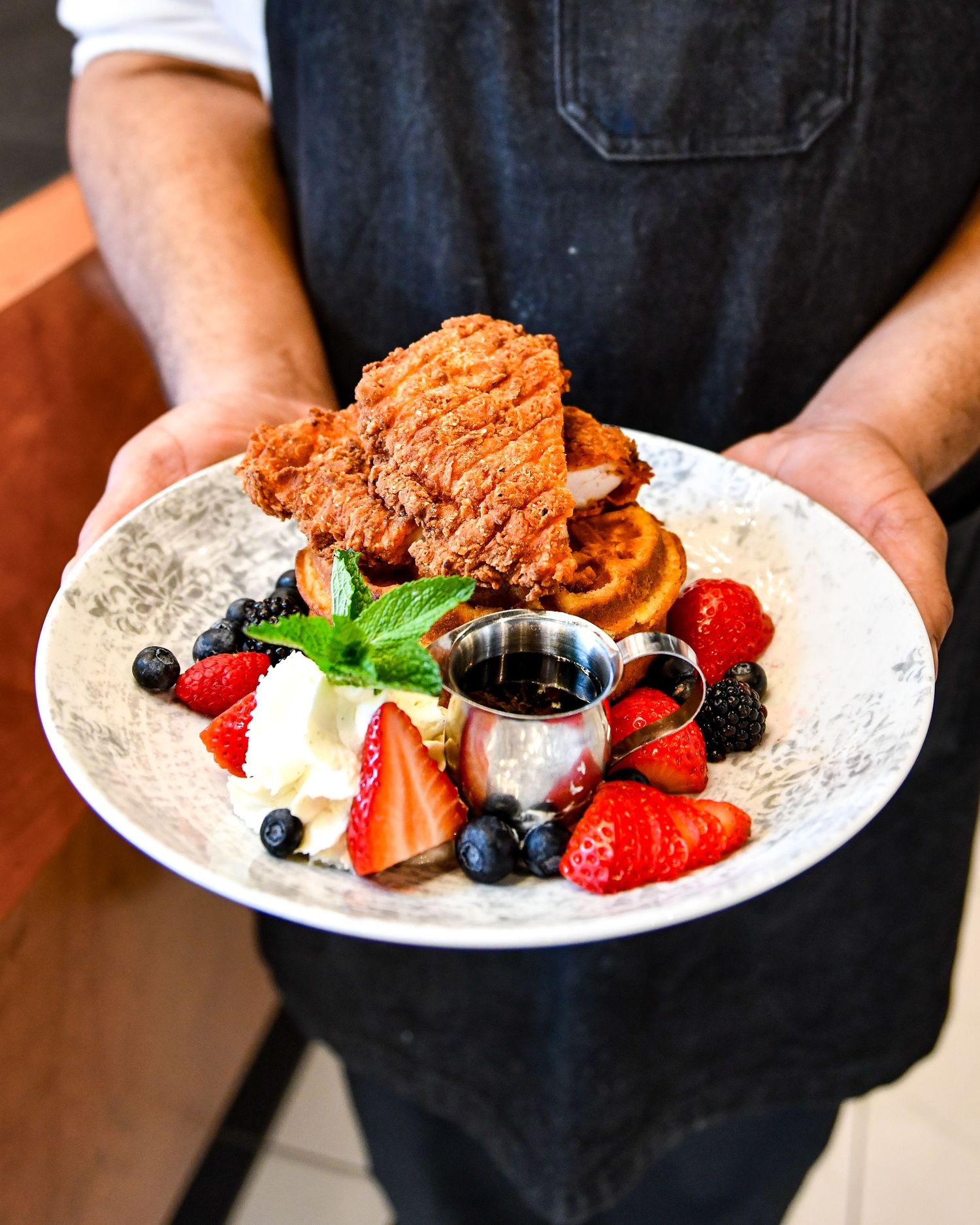 A plate of chicken and waffles garnished with fresh berries, mint, and served with syrup in a black apron presentation.