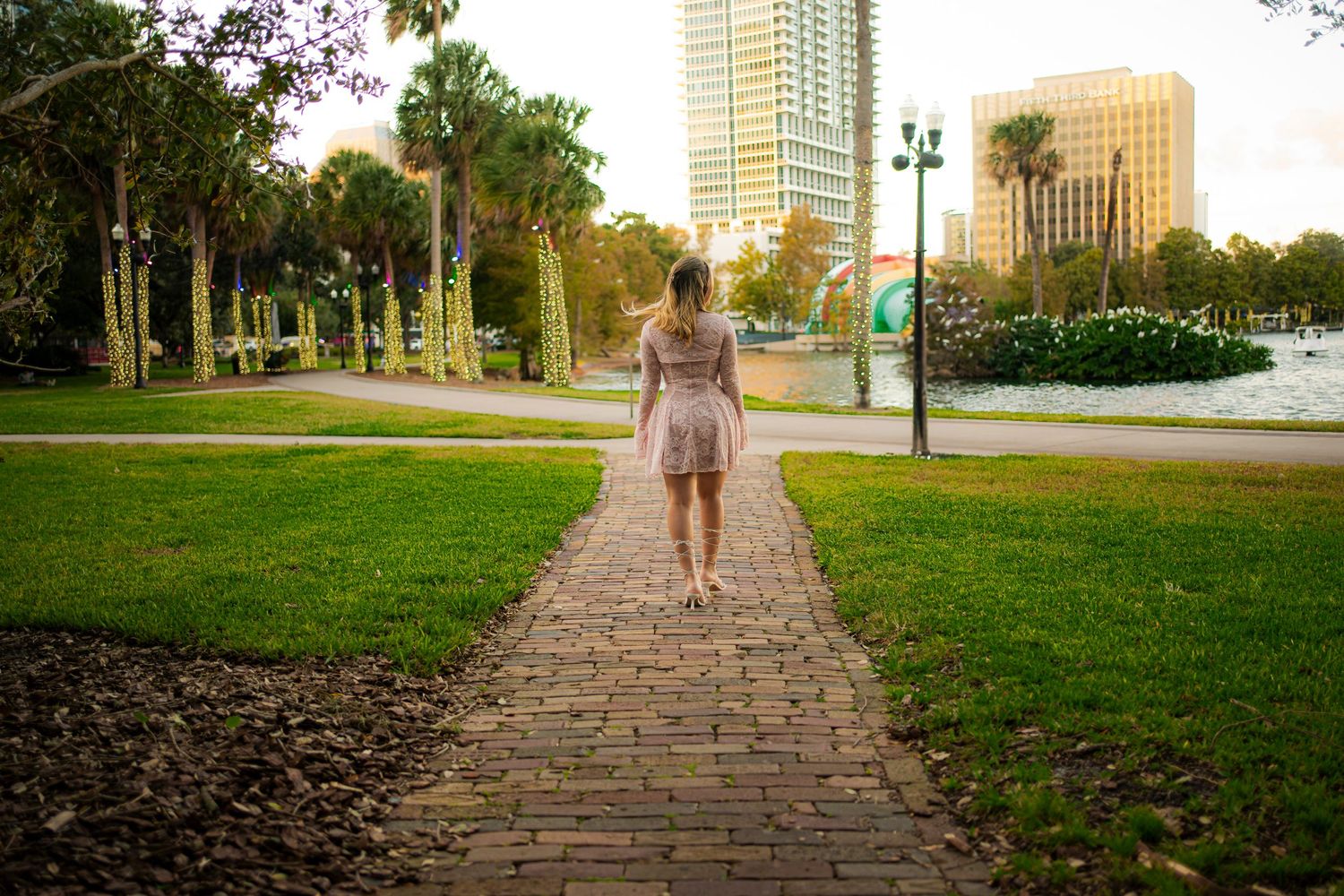 Figure walking alone on brick path through urban park with skyscrapers in background.