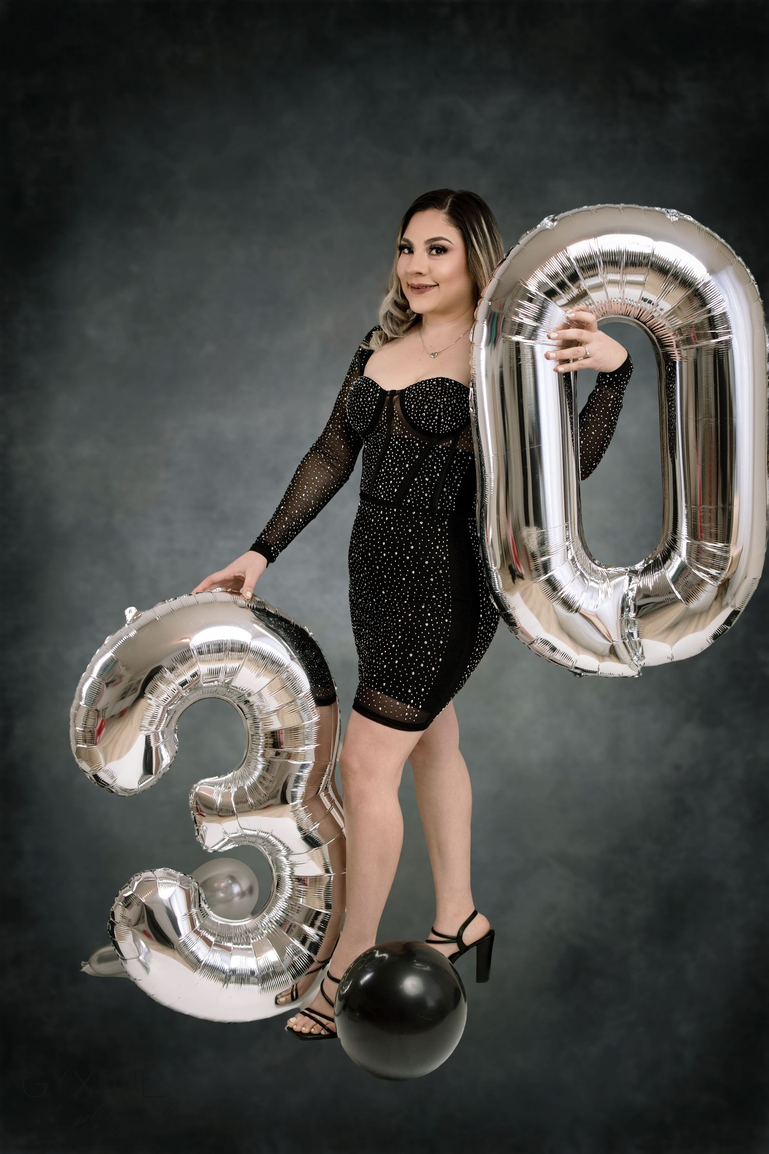 Celebratory portrait with silver 30th birthday balloons and black sequin dress against dark background.
