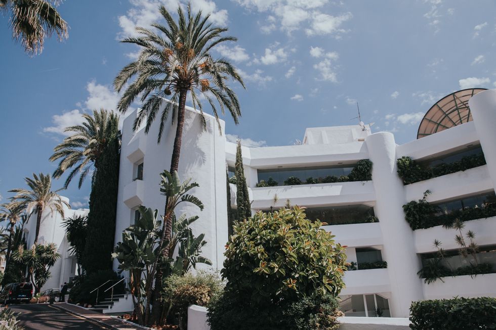 Mediterranean-style white apartment building with palm trees and blue sky in a coastal resort setting.