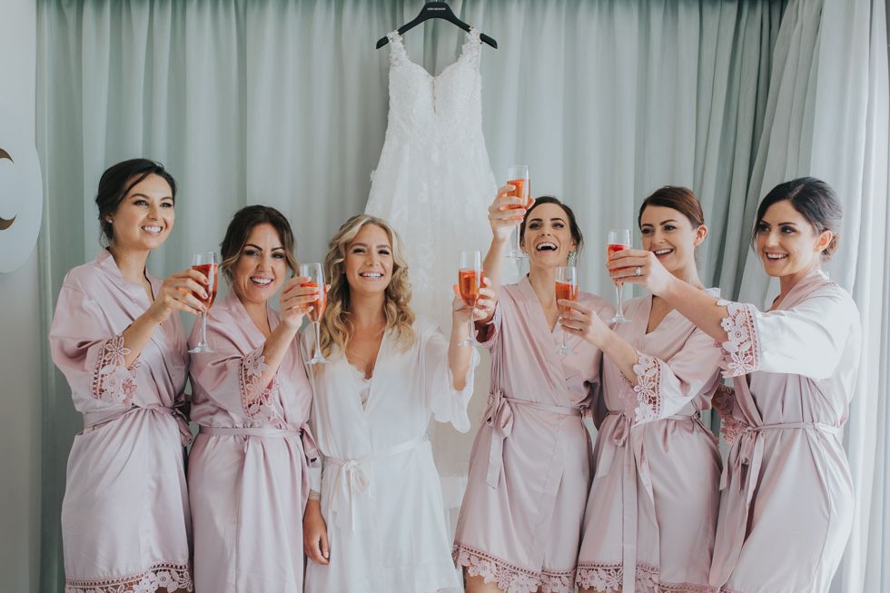 A wedding dress hangs above a group in matching pink satin robes raising champagne glasses in celebration.