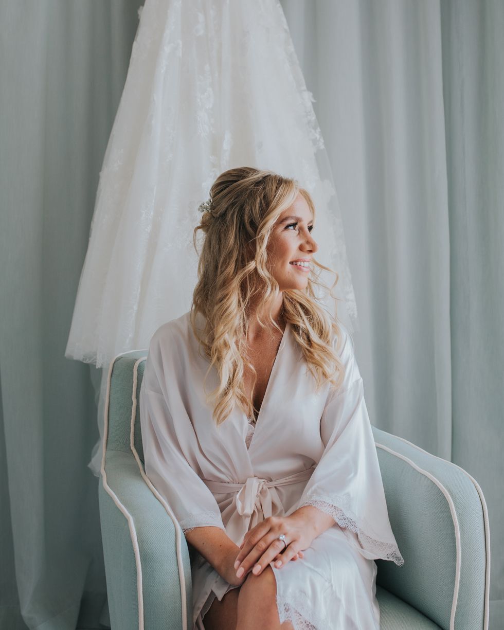 Elegant bridal photo with soft white robe against mint green curtains as wedding dress hangs in background.