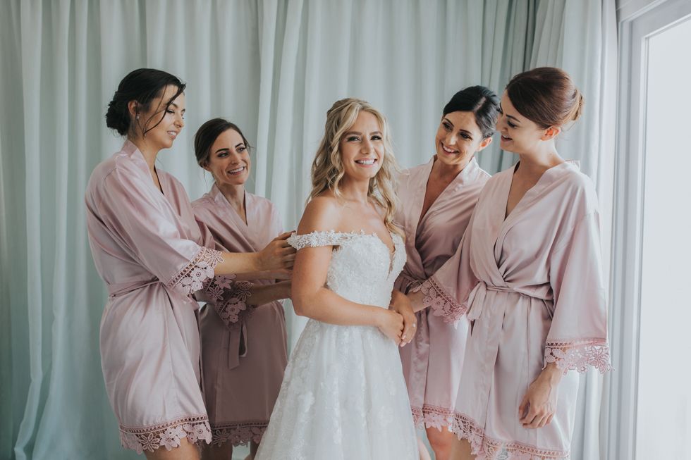 A bride in a white wedding dress stands with bridesmaids wearing blush pink robes while getting ready for the ceremony.