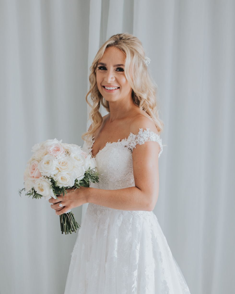 A bride in a lace wedding gown with off-shoulder details poses with a pink and white bouquet against white curtains.