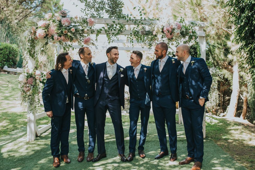 Wedding party in navy blue suits stand together laughing under floral arch in sunny garden setting.