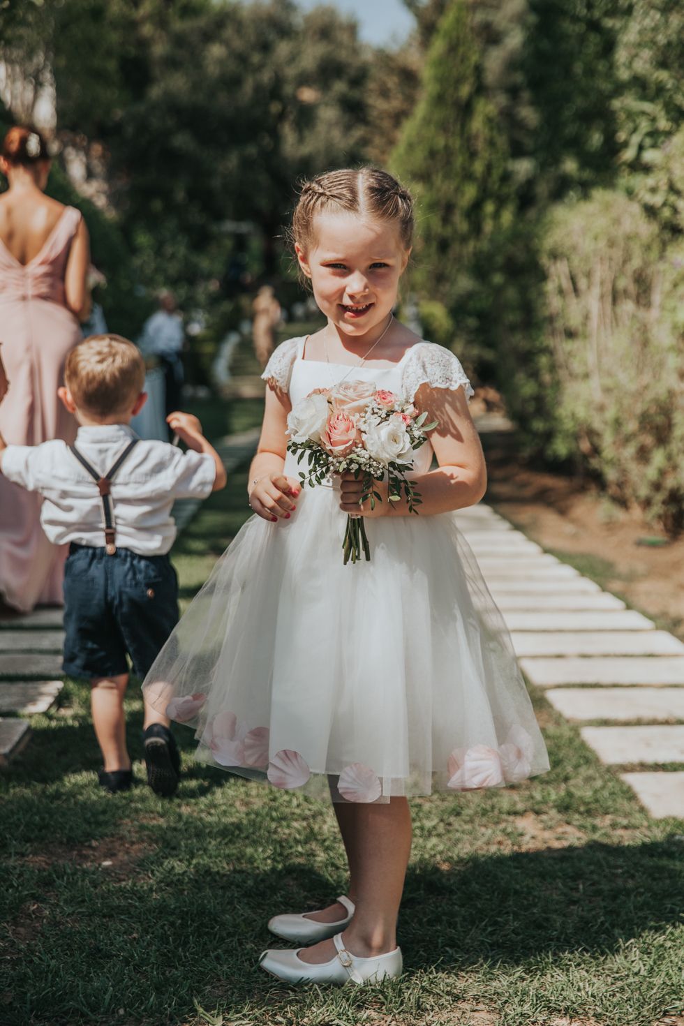 Young flower girl in white tulle dress holds a small bouquet while standing on a garden pathway.