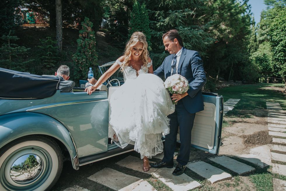 Couple exits vintage blue car on wedding day as bride in white dress steps out with groom's assistance in sunny garden.