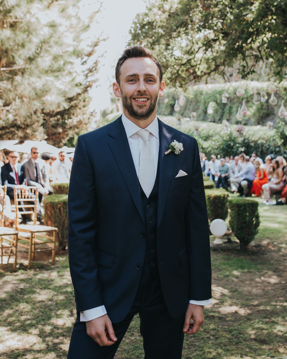 A person in a navy three-piece suit and cream tie smiles while standing outdoors at a wedding ceremony.