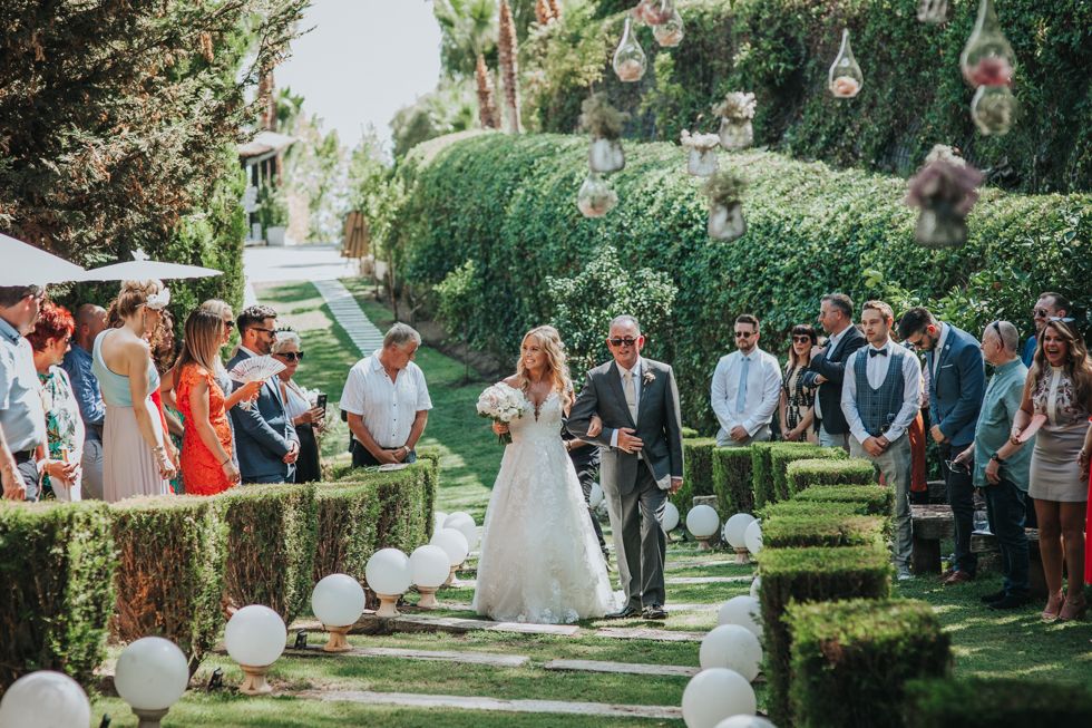 Outdoor garden wedding ceremony with white aisle balloons and hanging string lights amidst lush greenery.